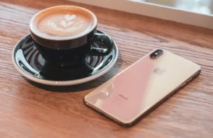 Mặt lưng iPhone XS Max màu vàng gold trên bàn gỗ, cạnh tách cà phê cappuccino