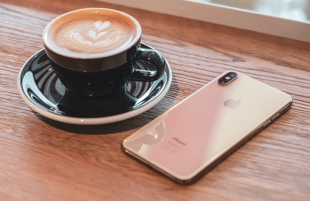 Mặt lưng iPhone XS Max màu vàng gold trên bàn gỗ, cạnh tách cà phê cappuccino